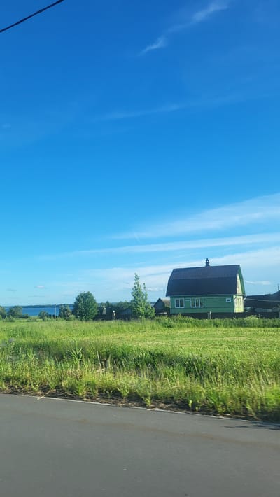 The image depicts a rural landscape with a clear blue sky and a few wispy clouds. In the mid-ground, a two-story house with a dark roof and light green siding stands in a grassy field. The house has several windows and a small chimney. Adjacent to the house, there are other structures and trees. A body of water, likely a lake or river, is visible in the far distance. The foreground shows a grey asphalt surface, possibly a road, with a strip of green grass and vegetation along its edge. The scene appears to be a sunny day in a village setting.