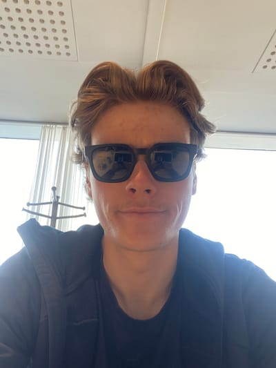 A close-up, slightly angled, indoor shot of a person wearing black sunglasses and a black jacket. The person has light skin and wavy, light brown hair. Behind them are a window with blinds and a white wall with a perforated panel. Reflected in the sunglasses are portions of the room's interior. The location is undetermined and there is no information to verify the location in Kastrup, Denmark.