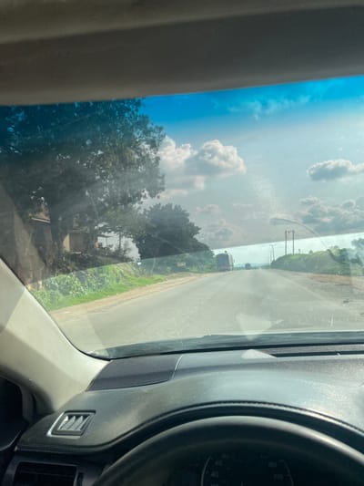 This image captures a view from inside a vehicle, looking through the windshield. The perspective is slightly angled, suggesting the vehicle is in motion or parked on an incline. Dominating the left side of the frame is the bright blue sky, adorned with fluffy white clouds, indicative of a sunny day. The sun's glare is visible on the windshield, creating a shimmering effect in the upper portion of the sky.

The scene outside consists of a paved road stretching into the distance, flanked by lush green vegetation and trees. In the mid-ground, utility poles with wires are visible, suggesting an inhabited area. There are also some structures, possibly buildings or vehicles, in the distance. The overall setting appears to be rural or semi-rural in Nigeria, given the location context. The time of day is likely daytime, given the bright sunlight. The weather appears to be fair and clear.

The interior of the vehicle is partially visible on the right, showing part of the dashboard, including a speaker grille and an air vent. A portion of the steering wheel is also visible, with a speedometer gauge faintly discernible, showing markings up to 200. There is no discernible text in the image. The image evokes a sense of travel or a journey through a sunlit landscape.