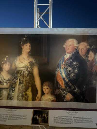 The image shows a framed reproduction of Francisco de Goya's painting "The Family of Charles IV." The painting depicts the Spanish royal family, including King Charles IV, his wife Maria Luisa of Parma, and their children and other relatives. The reproduction is displayed outdoors, with a blue sky and a metal structure visible in the background, suggesting it might be part of an exhibition or public display. Text panels with information about the painting and the artist are visible below the reproduction. The location is identified as Mazarrón, Spain, where a sign indicates "PINTURA ESPAÑOLA" (Spanish Painting). The reproduction is large, with a prominent gold frame. The painting itself is detailed, with the figures in formal attire, and Goya's characteristic use of light and shadow is evident.