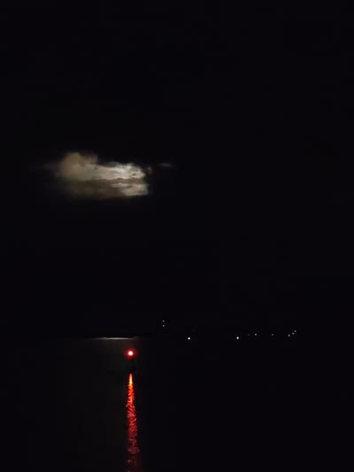 The image presents a dark, tranquil nighttime scene over a body of water in Rodino, Russia. The sky is largely black, with a patch of illuminated clouds visible in the upper left quadrant, suggesting moonlight filtering through them, though the moon itself is not directly visible.
The main focal point is a bright red light source positioned on the water, likely a buoy or a navigational marker. This red light casts a vivid, elongated reflection that stretches diagonally downwards and to the right across the dark, still surface of the water. In the far distance, several faint, tiny light points are discernible along the horizon, hinting at a distant shoreline or other structures.
The overall setting is one of profound darkness, with the few light sources providing a strong contrast. No people are visible, and there is no explicit activity taking place, conveying a sense of quiet solitude. The weather appears calm, as evidenced by the clear reflection on the water. No visible text is present in the image.