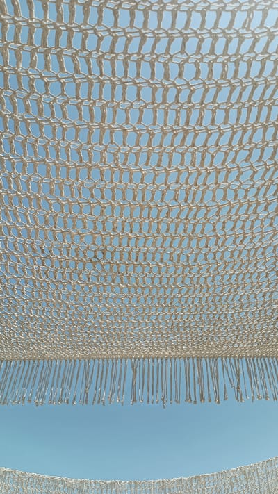 The image captures a bright, clear blue sky viewed through a textured, cream-colored woven net. The net is draped overhead, creating a layered effect. The upper portion of the image is dominated by the intricate pattern of the net, which is composed of numerous small, interlocking loops. This net has a fringe of hanging strands along its lower edge. Below this net, a wider expanse of the blue sky is visible, with another section of similar net appearing at the very bottom of the frame, also with hanging fringe. The overall impression is one of an outdoor setting, possibly a beach or a seaside promenade in Nea Peramos, Greece, where such decorative canopies are often used to provide shade. There are no people or specific objects visible other than the net and the sky.