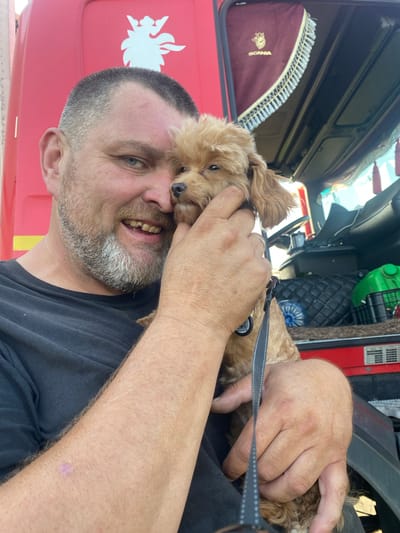 In Mais'ke, Ukraine, a man with a graying beard and short hair, wearing a black t-shirt, holds a small, light brown dog close to his face. The man is smiling broadly, with his teeth visible. He is holding the dog's head with his right hand, while his left arm is around the dog. The dog's leash is visible, with a tag attached. In the background, the red side of a Scania truck is visible, featuring the brand's logo. The interior of the truck's cabin is also visible, with dark upholstery, a steering wheel, and a green container. The scene appears to be outdoors, with daylight present.