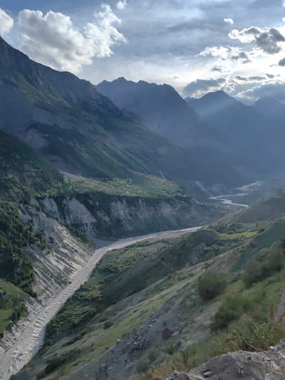 This vertical, full shot captures a wide, arid valley in Tiling, India, under a dramatic sky filled with dark, heavy clouds and shafts of sunlight. The valley is dominated by steep, sparsely vegetated slopes, with a wide, muddy river snaking through its center. The riverbed is a mix of light-colored sediment and flowing water, suggesting recent rainfall or snowmelt.

On the left side of the valley, the slopes are scarred by erosion, with lighter colored rock formations contrasting with the darker, more vegetated areas. Patches of green vegetation, including trees and shrubs, cling to the hillsides, particularly in the middle and upper portions of the valley. In the distance, smaller, more distant mountain ranges are visible, shrouded in haze and shadow, creating a sense of depth.

Near the top of the frame, scattered buildings, indicative of a small settlement, are nestled on a cultivated terrace. These structures appear small and insignificant against the vastness of the landscape. The sky is a dynamic mix of dark storm clouds and brighter areas where sunlight breaks through, casting rays that illuminate parts of the mountains. The overall impression is one of raw, untamed natural beauty and the vastness of the Himalayas.