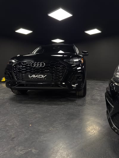 The image shows the front of a black Audi SUV parked indoors. The vehicle is the primary subject, with a focus on its grille and headlights. The background is dark, with four illuminated rectangular ceiling lights visible. The floor is a dark, textured surface that appears to be concrete or a similar industrial material, with some scuff marks and debris. To the right, the front of another black car is partially visible. The overall lighting suggests an indoor space, possibly a showroom or a detailing studio, with the bright ceiling lights creating reflections on the car's surface. There are no people visible in the image. The logo on the car's license plate area reads "LAKOV DETAILING". The context provided about the city of Giurgiu, Romania, is not visually apparent from the image itself.