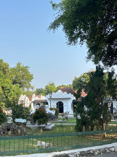 This image captures a view of the Taman Sari Gua Sunyaragi in Cirebon, Indonesia. The foreground features a green metal fence with decorative gold finials. Beyond the fence, a light-colored, possibly calico cat is walking on a grassy lawn. In the middle ground, the distinctive white, intricately decorated buildings of Taman Sari Gua Sunyaragi are visible, set amidst lush green trees. Some of these buildings feature ornate white sculptures, including what appear to be stylized animal figures. A lamppost with a globe-shaped light fixture stands near one of the buildings. The background consists of a clear blue sky and more foliage. The overall scene suggests a well-maintained historical and cultural site bathed in natural light.