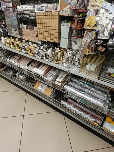 A shelf stocked with various home decor items. Decorative metallic apple-shaped objects in gold and silver line the top shelf. Below, wire baskets contain small wooden planks, craft supplies, and rolls of decorative paper with chevron patterns. Other items such as glass tiles, mosaic grids, leaves, and picture frames can be seen on the top shelf.