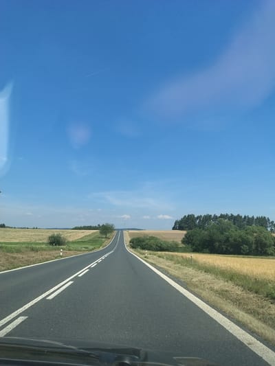 The image captures a view from a vehicle driving on a two-lane asphalt road that stretches into the distance, curving slightly to the right and uphill. The road is marked with solid white lines on both sides and a broken white line in the center. The sky above is a clear, bright blue with a faint contrail visible in the upper left quadrant and a soft, diffused cloud or light reflection in the upper right quadrant.

The landscape on either side of the road consists of fields. On the left, there are fields with dry grass or stubble, with some patches of green vegetation and a solitary tree. On the right, the fields are golden, suggesting crops like wheat or barley, with taller green bushes and trees bordering them. A line of dark green coniferous trees forms a dense background on the right side of the road in the mid-ground.

The terrain appears rural and agricultural. The overall impression is that of a sunny day in the Czech countryside, possibly near Horažďovická Lhota, given the context provided, though no specific landmarks within the city are identifiable from this perspective. The foreground shows the top of a car's dashboard, indicating the photograph was taken from the driver's or passenger's perspective.