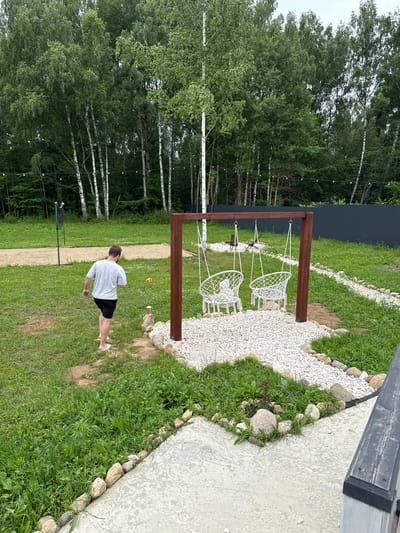 The image depicts an outdoor recreational area in Ryzhkovo, Russia. A man wearing a light-colored t-shirt and dark shorts is walking on a grassy lawn. In the background, a wooden swing set with two white macrame chairs is positioned over a gravel bed. Adjacent to the swing set, a dark metal fence encloses the property. A volleyball net and sandy court are visible further in the background, surrounded by a dense forest of birch trees. The ground is covered in grass, with some areas of bare soil and decorative gravel pathways. A small orange ball lies on the grass near the man. The sky is overcast.