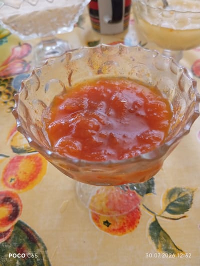 A close-up, eye-level shot shows a glass dessert dish filled with a vibrant orange jam or compote. The dish is ornate, with a scalloped rim and textured glass. The jam is chunky, with visible pieces of fruit. The dish sits on a tablecloth with a yellow background and a pattern of colorful fruits, possibly apples, and green leaves. In the background, out of focus, other dishes containing food items are visible, one appearing to hold a lighter-colored dessert. The lighting is soft and natural. The timestamp "30.07.2026 12:32" is visible in the bottom right corner, and "POCO C65" in the bottom left. No people or specific location within Beruniy, Uzbekistan are identifiable from the image.