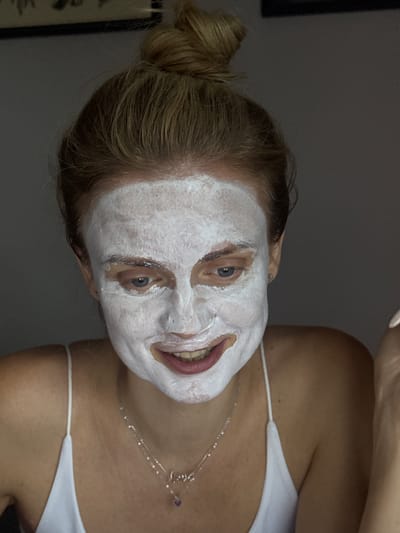 The image shows a close-up portrait of a young woman with fair skin and blonde hair pulled into a messy bun. She is wearing a white slip and a silver necklace with a small purple heart charm. Her face is covered in a white face mask, obscuring most of her features except for her eyes, nose, and mouth. She is looking downwards with a slight smile. The background is dark and out of focus, with hints of a picture frame on the upper left. The image does not provide any information about the location in the city of Лесные Поляны, Russia.