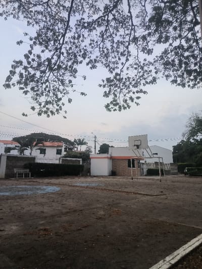 The image displays an outdoor recreational area in Cúcuta, Colombia, under an overcast sky partially obscured by the dark silhouettes of leafy tree branches in the foreground. The ground consists of unpaved, compacted dirt with faint, faded blue and dark line markings from a former court surface. A white concrete curb defines the bottom right edge.

In the midground, a metal basketball hoop with a white rectangular backboard and black rim stands to the right, adjacent to a white metal soccer goal frame lacking a net. Behind these structures, a single-story building is visible, featuring a white facade, an orange horizontal strip, and a section clad in brown brick-patterned material, with a dark-tinted window. To the left of this building, a dense green hedge borders a low wall, in front of which a white park bench rests next to a gray block wall.

The background reveals several white residential buildings with distinctive red-tiled roofs, characteristic of the region. Various trees, including tall palm trees and a large, broad-canopied tree, are interspersed among the buildings. Overhead utility lines traverse the upper midground horizontally. No individuals are present in the scene.