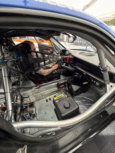 The image shows the interior of a racing car from the rear passenger side. The driver, a man with tattoos, is wearing a black and white racing suit and sunglasses. He is sitting in the driver's seat with his right hand on the steering wheel and his left hand on the gear shifter. He is looking straight ahead. The car has a roll cage, a racing seat, and a steering wheel. The car is a black and white Porsche 911 GT3 R. The location is Stavelot, Belgium, at the coordinates 50.441, 5.969. The environment is a racing track. The car is in the pits. The image is taken from the back of the car. The camera is pointed towards the front of the car.