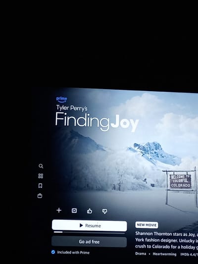 The image displays a photograph of a digital screen, likely a television, captured at a slight angle in a dimly lit indoor environment. The screen is presenting content from Amazon Prime Video.
The central subject on the screen is the title card for "Tyler Perry's Finding Joy," with the Amazon Prime logo positioned above the title. The background image for this title features a wide, panoramic view of a snowy, mountainous landscape under an overcast sky. Several snow-covered evergreen trees are visible, and a rustic wooden sign on the lower right reads "WELCOME TO COLORFUL COLORADO."
Various user interface elements are present around the title and background. On the far left, a vertical navigation bar displays icons for search, a grid-view (browse), a bookmark (watchlist), and a shopping cart. Below the central image, a horizontal row of icons includes a plus symbol (add), a film reel (share), a thumbs-up (like), and a thumbs-down (dislike). A prominent white button with a play symbol is labeled "Resume," situated above a darker button stating "Go ad free." Below these, a blue checkmark indicates "Included with Prime."
To the lower right, additional information about the movie is displayed, including a "NEW MOVIE" tag. Partially visible text describes the plot: "Shannon Thornton stars as Joy, a York fashion designer. Unlucky in crush to Colorado for a holiday g". Genre classifications "Drama" and "Heartwarming" are listed, along with an "IMDb 4.4/10" rating.
The image itself contains no specific visual cues to identify its physical location in Royton, United Kingdom. The content depicted on the screen refers to a fictional setting in "Colorado," not Royton, UK.
