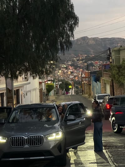 This image captures an urban street scene at twilight in Guanajuato City, Mexico. In the foreground, a dark grey BMW X3 SUV is stationary on a wet asphalt street, facing the viewer at a slight angle, with its headlights active. The front passenger door is open. A female occupant is seated inside, smiling and gesturing with her raised right hand towards the camera. A second female subject is partially depicted exiting the open passenger door, wearing a dark jacket and blue wide-leg jeans, with her right leg visible on the pavement. A third female subject stands immediately to the right of the vehicle, also in a dark jacket and blue jeans, facing towards the SUV. The street, lined with residential structures, descends into a valley. The midground and background reveal a densely packed cityscape of colorful buildings, illuminated by numerous streetlights and interior lights, extending up surrounding hillsides. A rugged mountain range is visible on the distant horizon beneath a pale, overcast sky. Utility lines are present overhead. Additional vehicles, including a white car and a dark pickup truck, are parked or stopped further down the street. A large tree with dark, dense foliage partially obstructs the upper left portion of the frame.