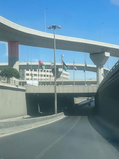 The image displays a multi-level road interchange in Amman, Jordan, identifiable as the Fifth Circle (Dawwar Al Khames) or a similar major urban junction. The scene is set under a clear blue sky during daylight hours.
In the foreground, an asphalt road leads into a dark, concrete underpass or tunnel. Concrete barriers line the road edges, and faint outlines of vehicles are visible within the tunnel's entrance. A tall, gray metallic street light pole, equipped with multiple floodlights, stands centrally on a median, extending upwards past an intermediate elevated concrete highway.
Between the lowest and intermediate road levels, a row of six flagpoles supports alternating flags: three Jordanian flags (black, white, and green horizontal stripes with a red triangle containing a white seven-pointed star) and three Palestinian flags (black, white, and green horizontal stripes with a red triangle).
In the background, behind the flagpoles, a multi-story building with a light-colored facade and numerous rectangular windows is visible. Several cellular antennae or communication towers are positioned on or near its roof. The scene is framed by multiple elevated concrete highways, supported by various concrete pillars. One cylindrical pillar on the left has a reddish-pink base. Another large, gray concrete support structure for an elevated road is visible on the right, adjacent to a blue square road sign with a white U-turn arrow. A small, bushy green tree is situated on a raised concrete planter on the left side of the frame. No individuals are discernible within the image.