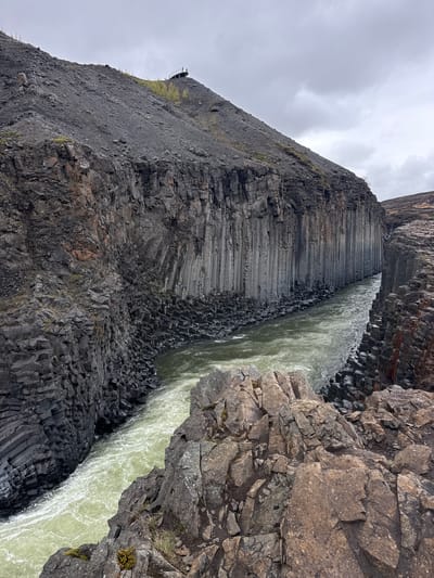 This media file presents a dramatic natural landscape, likely a canyon or gorge, dominated by distinctive geological formations. The central feature is a river with turbulent, light green water flowing between towering, dark cliffs.

The most prominent geological formations are the extensive basalt columns that form the canyon walls, particularly visible on the left side of the river. These columns are dark grey to black, exhibiting precise, often hexagonal or polygonal shapes, stacked vertically and horizontally, creating intricate geometric patterns. Some sections appear like giant steps formed by horizontal columns. The right side of the canyon displays more irregularly fractured and rugged rock formations, also dark and stony.

The river itself is a striking pale green, indicative of mineral content or glacial meltwater, and is actively flowing with visible white foam and ripples. The water appears powerful as it courses through the narrow gorge.

The sky above is overcast with soft, grey clouds, suggesting a cloudy daytime. No direct sunlight is visible, contributing to a muted, natural color palette. High on the cliff edge on the left, a small wooden structure, possibly a viewing platform with railings, can be discerned, indicating the area is accessible to visitors. There are no people visible in the frame, nor any discernible text. The scene captures the raw, powerful beauty of a volcanic landscape, consistent with the specified context of Iceland.