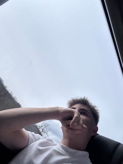 A young adult male is depicted from a low-angle perspective, seated within a vehicle. He wears a plain white short-sleeved t-shirt. His head is tilted upwards, with his eyes directed towards the upper frame. His right forearm is raised, with the hand positioned near his mouth; specific finger arrangement shows digits curled. The individual's hair is short, light brown, and appears disheveled. To the right, a dark headrest or seat portion of the vehicle is visible. The upper two-thirds of the image display an overcast, light grey sky visible through a vehicle window. A section of a distant, denuded hillside featuring bare trees is visible through the left side of the vehicle window. The scene is located in the city of Dalbok Dol, Bulgaria.