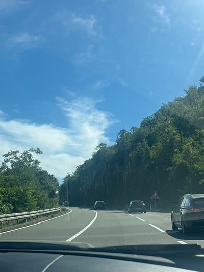 The image depicts a view from inside a vehicle driving on a winding, paved road in Donji Ulići, Montenegro. The road has clear white lane markings. On the left side of the road, a white guardrail runs along the edge, separating the road from a steep, tree-covered embankment. The right side of the road is bordered by a rocky cliff face densely covered with green foliage. Several cars are visible on the road: one dark-colored station wagon in the foreground right, another dark car further ahead, and a third car in the distance to the left. A triangular warning sign with a red border and a symbol indicating falling rocks is posted on a pole to the right of the road. The sky is a bright blue with scattered white clouds. The sun is shining, casting shadows on the road and vegetation. The scene appears to be a rural or mountainous area.