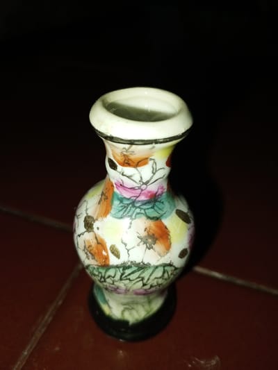 A small, decorative ceramic vase is depicted standing on a dark reddish-brown tiled surface in Longmiang, Laos. The vase measures approximately 7-10 cm in height and features a narrow neck with a flared rim, a wider bulbous body, and a tapered base. The rim and base are outlined in dark grey or black. The body of the vase is white with a multicolored floral pattern, including prominent orange-brown, pink, and teal/green flowers or leaves, accented with small, irregular gold-colored spots. The background is predominantly dark, suggesting low ambient lighting or a shadowed area, with direct illumination on the vase from the front or upper-front. No individuals, actions, or interactions are visible.