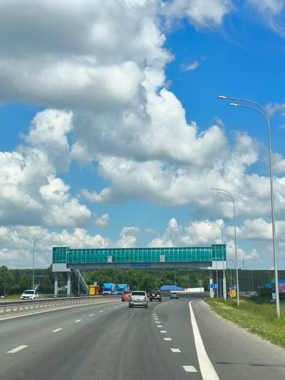 This is a medium-distance, eye-level, exterior, vertical shot taken from a vehicle on a highway in Shikhazany, Russia. The highway features multiple lanes and is bordered by a metal guardrail and a grassy shoulder. In the foreground, several cars are visible, driving in the same direction. A large pedestrian bridge with green tinted glass spans the highway in the middle ground. Beyond the bridge, a treeline and rolling hills are visible under a bright blue sky filled with large, puffy white clouds. Streetlights and road signs are also present.