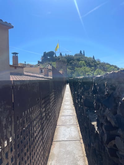 The image shows a view from the top of the medieval wall in Hostalric, Spain. The perspective is along the length of the fortified wall, with a walkway made of rectangular slabs running between the rough stone inner wall and a metal grate parapet on the outer edge. Beyond the wall, there are tiled rooftops and other buildings. The sky is clear blue. A yellow flag is visible on a tower in the distance. Trees and vegetation cover the landscape beyond the town.
