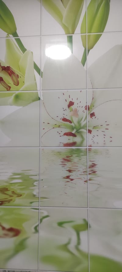 This is a close-up, eye-level shot of a tiled wall featuring a floral design with lily flowers and reflections on water. The tiles are arranged in a grid pattern. The upper portion of the image shows parts of several lily flowers, with white petals and green stems and buds. Reddish-brown stamen are visible in the center of one flower. The lower portion of the image depicts a wavy, rippled reflection of the flowers on what appears to be water. The overall color palette consists of whites, light greens, and hints of reddish-brown. The image is devoid of any human presence or activity. The scene does not provide any contextual information to determine its location within the city of Ridder, Kazakhstan.