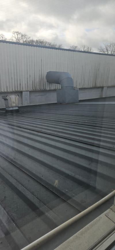 The image presents an elevated, oblique view of a section of a corrugated metal roof, likely belonging to an industrial or commercial building. An L-shaped metal duct with a square base is mounted on the roof. A smaller, box-like structure is attached below it. Patches of darker residue or staining are visible on the roof's surface near the ductwork. The background shows a hazy sky, potentially suggesting overcast weather. The presence of bare tree branches along the roof's edge implies a winter season. The image is seemingly taken from inside a building, looking out through a screen, as evidenced by the mesh pattern overlaying the scene. The scene is located in Delbrück, Germany.