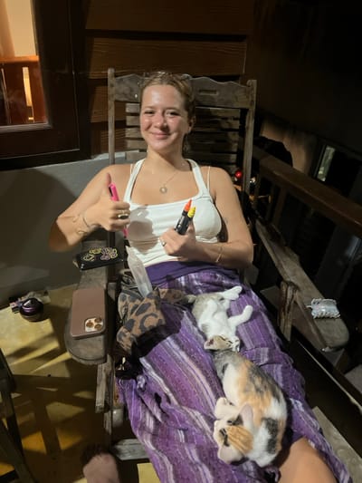 A young adult female is seated in a wooden rocking chair in Baan Nai Wok, Thailand, at night. She has light brown hair, is smiling, and wears a white tank top and a purple patterned sarong. Her left hand is raised in a thumbs-up gesture, holding a pink marker. Her right hand holds two additional markers, one orange and one yellow. On her lap, two kittens are sleeping on the purple fabric; one is a calico, and the other is light gray and white, both lying on their backs or sides.

To her left, on the armrest of the chair, rests a brown smartphone with multiple camera lenses and a black case featuring a yellow abstract design. Below these items, a small, dark, lidded container is visible. A brown and black leopard-print bag is situated on her lap next to the phone. The background consists of a dark wooden wall to the left, with a window showing a dark exterior, and a wooden railing to the right, suggesting an outdoor or semi-outdoor porch area. The scene is illuminated by an unseen light source.