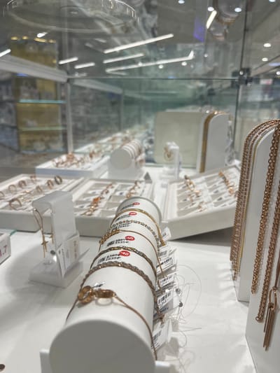 This image shows a display of jewelry, likely in a store, with a focus on bracelets and necklaces. The jewelry appears to be made of gold or rose gold, with various chain styles and decorative elements like beads and rings. Some items have price tags attached, indicating they are for sale. The display is organized on white stands and trays, with a glass case in the foreground and background suggesting a retail environment. The lighting is bright, typical of a store. No people are visible in the image. The presence of Russian price tags and Cyrillic text on the labels suggest the location is in Russia. The city of Kokkorevo, Russia is a possible location for such a store.