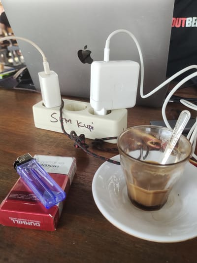 A flat lay captures a moment of relaxation on a wooden table. The scene is dominated by various objects, suggesting a break or downtime. A laptop, partially visible in the background, is plugged into a power strip with two white Apple charging bricks. The power strip has "SM kopi" written on it in purple marker, indicating "coffee" in Indonesian.
In the foreground, a pack of Dunhill cigarettes with a purple lighter placed on top is positioned to the left. The cigarette pack displays text like "SMOKERS DIE YOUNGER" and "DUNHILL TOBACCO LONDON LIMITED". To the right, a small glass of coffee sits on a saucer, with a spoon resting inside. The coffee appears to be a light brown color, with a visible layer of froth at the top. The overall lighting suggests it might be daytime, and the setting seems to be indoors, possibly a cafe or a home, given the presence of a laptop and charging devices. There are no people visible in the frame, but the arrangement of objects implies someone has recently been using them or is about to. The mood conveyed is one of leisure and possibly contemplation.