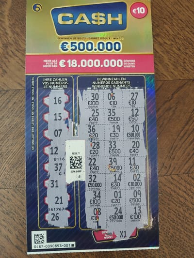 The image shows a lottery ticket with several numbers scratched off. The lottery ticket has the word "CASH" prominently displayed on it, along with monetary values in Euros. There are multiple rows and columns of numbers, some of which are partially obscured by scratch-off material. Some of the text on the ticket is in Dutch and French, indicating it is likely from Belgium. There is a QR code present, labeled "RESULT?" and "SCAN IN APP." The background of the image appears to be a wooden surface. The lottery ticket is positioned on this surface, slightly angled. The lighting suggests it is an indoor setting, possibly a home or a shop. There are no people visible in the image. The overall impression is of a gambling or lottery-related item presented for examination. This lottery ticket is not located in Molenbeek-Saint-Jean - Sint-Jans-Molenbeek, Belgium; its location is unknown and cannot be determined from the image.