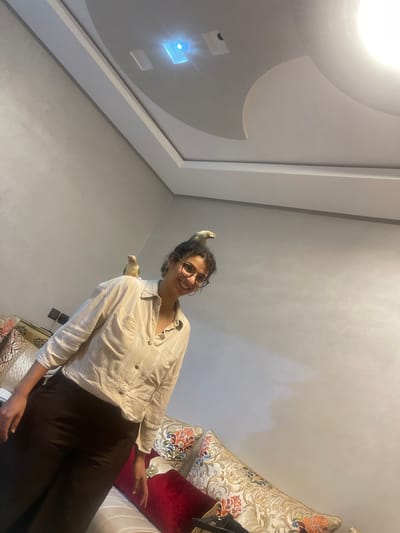 The image shows a woman with curly dark hair and glasses, smiling and looking upwards. She is wearing a light-colored button-up shirt and dark pants. She has two birds on her, one perched on top of her head and the other on her shoulder. She stands near a couch that has several decorative pillows. The interior appears to be inside a residence in Ait Ourir, Morocco, based on the style of the wall and pillows. The ceiling has a modern, recessed lighting fixture.