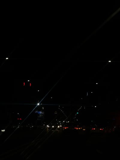 The scene is a nighttime view of a city street in Beni Mellal, Morocco, characterized by extremely low light conditions and reliance on artificial illumination. The foreground and mid-ground depict a multi-lane roadway with active vehicular traffic, primarily identified by multiple sets of white headlights and red taillights. To the upper left, two red pedestrian signals displaying the "don't walk" figure are visible. In the mid-ground, a green traffic light is observable on the right side of the road, and a discernible McDonald's "M" logo is present on a structure or sign. Various streetlights and other ambient light sources create points of light throughout the dark environment. No individuals are clearly visible. Prominent lens flares, appearing as diagonal streaks, traverse the image.