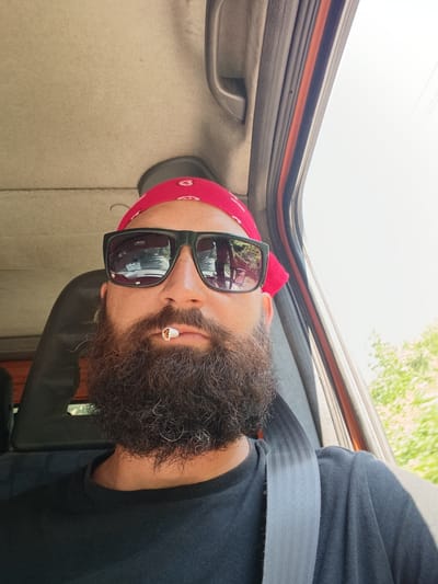 In Kresna, Bulgaria, a man with a long, dark beard and a red bandana is seen from the chest up, seated in a vehicle. He is wearing black sunglasses, and a cigarette is held between his lips. The man has a seatbelt across his chest. The interior of the vehicle is visible, including the headrest of a seat and the roof lining. Through the open car door, greenery and trees are visible, suggesting an outdoor environment, possibly a road. The lighting suggests it is daytime.