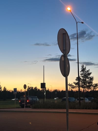 The image depicts a street scene at dusk in the city of Vyazovka, Russia. The sky transitions from a pale orange at the horizon to a deep blue overhead, with scattered clouds. A street lamp with an illuminated orange bulb is visible on the right side of the frame, casting a glow. Two round traffic signs are mounted on a pole in the foreground, facing left. In the middle ground, a dark-colored minivan is parked, with its taillights illuminated. To the left of the minivan, a white car is driving on a grassy area. A row of trees forms a dark silhouette against the sky in the background. The foreground consists of an asphalt road and a sidewalk. There are no people visible in the image.