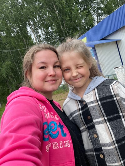 A selfie is taken outdoors in what appears to be a rural or semi-rural area, possibly near the city of Baldeyka, Russia, as indicated by the presence of birch trees, a simple building with blue corrugated metal siding, and what could be an outdoor utility structure.
In the foreground, two individuals are captured. On the left is an adult female with blonde hair pulled back, wearing a bright pink hooded sweatshirt with blue lettering. She is smiling and looking directly at the camera. Her nose piercing is visible. To her right is a young girl with blonde hair, also looking towards the camera and winking. She is wearing a grey hooded shirt layered under a black and white plaid jacket. The two are standing close together, suggesting a familiar relationship.
The background consists of a cluster of birch trees with green foliage against a cloudy sky. Behind them is a building with white siding and a prominent blue corrugated metal roof and trim. To the right of the building, a white cylindrical object, possibly a large bag or container, is partially visible. The ground appears to be dirt or gravel. The overall impression is casual and outdoor.