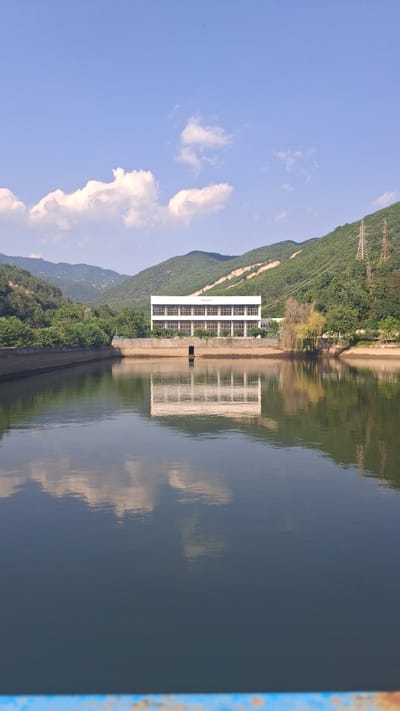 The image depicts a serene landscape dominated by a large, calm body of water reflecting the surrounding environment under a clear blue sky with scattered white clouds. A long, rectangular building with numerous windows stands prominently on the far bank of the water, its facade also reflected in the water. The building appears to be a dam control or administrative structure, likely part of the Gorno Spanchevo Dam in Bulgaria, given the context. Green, tree-covered hills rise behind the building, forming a natural backdrop. The water's edge is lined with a concrete embankment. The overall scene is one of natural beauty and functional infrastructure coexisting harmoniously.