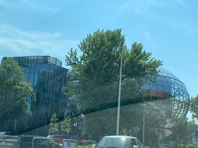 The media file captures an exterior view of a modern, architecturally unique building complex under a clear blue sky with scattered clouds, suggesting daytime. The primary subjects are the buildings, characterized by their extensive use of glass and distinctive geometric designs. One building features a faceted, almost crystalline facade, while another part of the complex is a large, spherical structure covered in a grid of glass panels, resembling a globe. Lush green trees surround and partially obscure the buildings, indicating a natural, possibly park-like setting.
In the foreground, several cars are visible, suggesting the scene is viewed from a street or parking area. The presence of cars implies that the buildings are accessible to the public, possibly serving as offices, commercial spaces, or a public institution. A lamppost stands prominently in the middle ground, further contributing to the urban or semi-urban context.
There is visible text on a sign in the lower left corner, which reads "ETY PARKINGOWE" and "OD 90 ZŁ/MIESIĄC". Another sign in the lower right corner has Polish text, including "TU ZAPARKUJESZ NA GODZINY" and "24/7 PLACE ZAPRASZA", indicating parking information and availability. These text elements, combined with the Polish language, strongly suggest the location is in Poland, and specifically Bielsko-Biała as per the provided context.
The overall impression is one of a bright, sunny day, highlighting the reflective surfaces of the buildings and the vibrant green of the foliage. No specific activity or event is overtly depicted, but the scene suggests a typical day in a developed urban environment. The clean lines of the architecture juxtaposed with the natural elements create a visually interesting and modern aesthetic. The image conveys a sense of a functional and aesthetically pleasing public space.