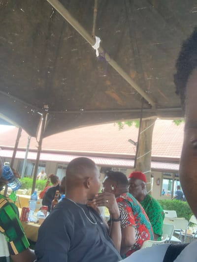 The image depicts an outdoor social gathering in Ogbunike, Nigeria, observed from a low angle under a dark fabric canopy supported by metal poles. The right side of a person's head, featuring dark, curly hair, occupies the immediate foreground. Several individuals are seated at tables in the mid-ground. From left to right, a person in a green and black patterned shirt holds a clear plastic water bottle. Next, a person with a short haircut wears a black short-sleeved top and a silver chain, with their left hand near their chin. Adjacent to them, a person with dark hair wears a brightly patterned multi-colored top and a ring on their left ring finger, with their right hand also near their chin. Further right, a man with a white beard wears a red cap and a green patterned top. The tables are laden with plastic water bottles, soda cans, and brown glass bottles. White plastic chairs are visible. In the background, beyond the tent structure and intervening foliage, stands a building with a reddish-brown tiled roof and multiple windows. A white fabric strip and a purple cord are tied around a vertical support pole of the canopy.