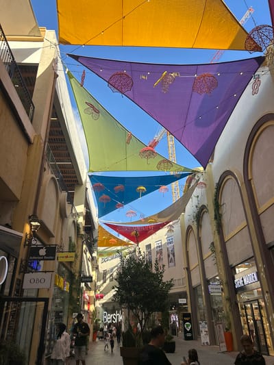 The image depicts a street scene in Hadera, Israel, characterized by an overhead canopy of colorful, triangular fabric shades in yellow, purple, green, and blue. Decorative wireframe umbrellas and other hanging ornaments are suspended between the shades. The architecture features light-colored buildings with arched windows and balconies. Several shops are visible, including "Steve Madden," "Bershka," and "OBO," along with various signs in Hebrew. A few people are walking along the street, including a woman in a white shirt, a man holding a drink, and a child. In the background, construction cranes are visible, suggesting ongoing development in the area. The overall atmosphere appears to be a pedestrian shopping area under a clear sky.