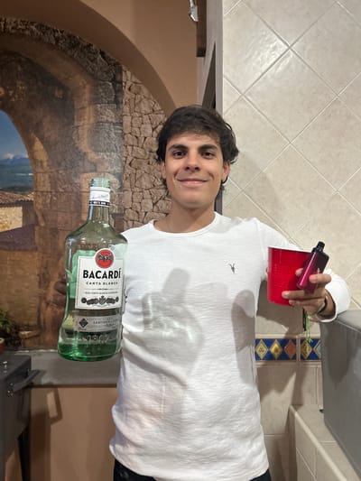 A person is holding a bottle of Bacardi Carta Blanca rum, a red plastic cup with a vaping device resting on top of it. The person is indoors. The background includes a decorative mural depicting a stone archway. The location is San Juan Cuautlancingo, Mexico.