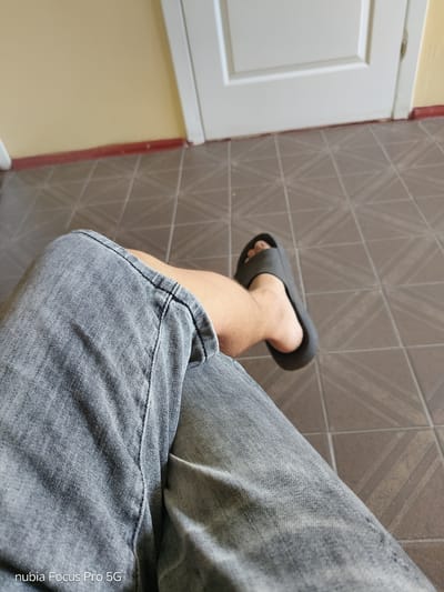 A close-up shot focuses on a person's legs, clad in light grey denim jeans. One leg is bent at the knee, with a black sandal on the foot. The background shows a tiled floor with a geometric pattern, and a glimpse of a closed white door against a yellow wall. The scene appears to be indoors, possibly in a residential setting. No specific landmarks or indications of San Martín de Porres, Peru are visible.