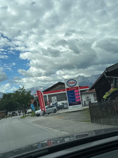 The image shows a gas station in Farchant, Germany, under a cloudy sky with mountains in the background. The prominent ESSO sign displays fuel prices: Diesel at 1.869, Synergy at 1.989, Supreme at 1.969, and Super E10 at 1.909. A red ESSO flag with the text "Willkommen bei Esso" is planted near the pumps. Several cars are visible, including a silver electric vehicle and a tractor to the right of the frame. The gas station appears to be part of the EG Group. The overall environment suggests a rural or semi-rural setting.