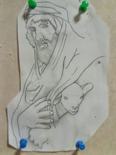 A pencil drawing of a bearded man wearing a head covering is pinned to a textured wall with three pushpins: one green pushpin at the top, a green pushpin on the upper right, and two blue pushpins at the bottom. The man is holding a lamb, which is nestled in his arms. The drawing is rendered in a simple, sketch-like style, with varying line weights and some shading to suggest form and texture. The background of the drawing is plain white paper. The wall appears to be a light beige or cream color with a subtle woven pattern. The drawing is positioned slightly off-center on the wall. This scene is located in Martuni, Armenia.