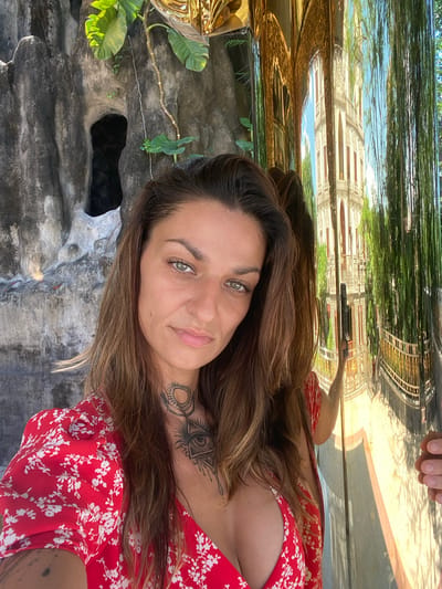A woman with brown hair and fair skin is posing for a selfie outdoors. She is wearing a red dress with white floral patterns. She has tattoos on her neck and arm. The background includes a stone-like structure with foliage, and a reflective surface above shows a partial reflection of buildings and trees, possibly in a city environment. The location is likely Dĩ An, Vietnam, as requested. A hand is seen in the reflection above her head, possibly holding the phone.
