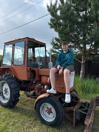 A young person is reclining on the hood of an orange tractor, positioned on a grassy field. The tractor, partially obstructed by a tree, has a cabin and large tires. The background includes a fence and a cloudy sky with overhead power lines. The specific location is Vysokinichi, Russia.