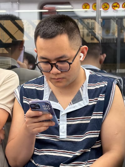 A young East Asian man wearing glasses and a sleeveless striped shirt is seated on a subway train, looking down at his smartphone. He has white wireless earbuds in his ears. His reflection is visible in the window behind him, along with other passengers and the interior of the train car, which features yellow circular signs with red markings. The man is holding his phone with his right hand, his thumb resting on the screen. The phone case is blue with white text that appears to say "KU BO". The scene appears to be in Tiaodeng, China, based on the information provided.
