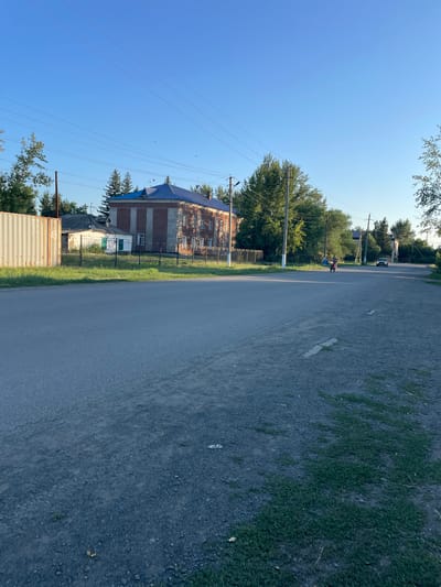 The image shows a street scene in a rural area, possibly the village of Kishkene-Kol in Kazakhstan. On the left, a large, beige shipping container is partially visible. Behind it, a low white building with a green door is fenced off. Further back, a large, older, brick building with a blue roof is prominent. Trees are scattered throughout the scene, with a line of taller trees visible behind the brick building. A paved road extends into the distance, with a person on a motorcycle in the middle ground and a car in the far distance. The sky is clear and blue, suggesting a sunny day. The overall impression is one of a quiet, possibly semi-abandoned, rural settlement.