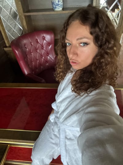 A woman with curly brown hair and blue eyes is wearing a white robe. She is positioned next to a red chair with a tufted design. The chair is located near a wooden shelf structure. Behind the woman, there is a red surface with a gold trim. The image does not provide enough information to verify the location in Cat Cat Village, Vietnam.
